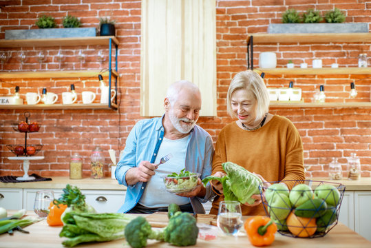 Cheerful senior couple eating salad standing together with healthy food on the kitchen at home. Concept of healthy nutrition in older age