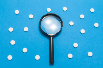 Black magnifying glass is on blue background surrounded by white pills as ornament polka dots....