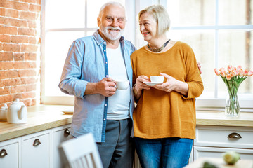 Fototapeta premium Portrait of a lovely senior couple talking together, standing with coffee cups near the window at home