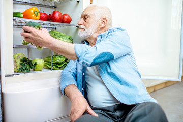 Handsome senior man taking fresh cabbage sitting near the refrigerator full of healthy products at home © rh2010