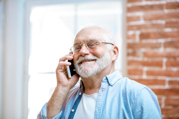 Cheerful senior grandfather talking with phone near the window at home