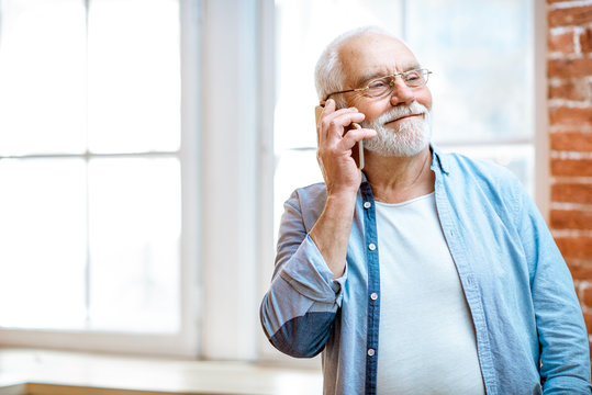 Cheerful senior grandfather talking with phone near the window at home