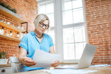 Beautiful senior woman working with documetns and laptop on the kitchen at home