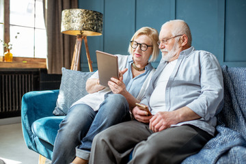 Lovely senior couple dressed casually using digital tablet while sitting together on the comfortable couch at home - obrazy, fototapety, plakaty