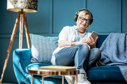 Senior woman dressed casually listening to the music with headphones and phone on the couch at home