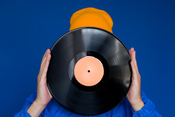 young man in a blue sport 90s style jacket and yellow hat holding vinyl record, man hides his face...