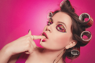 Cheeky girl with bubble gum posing on pink background in body, with curlers on head. Pretty woman with sweet makeup in studio