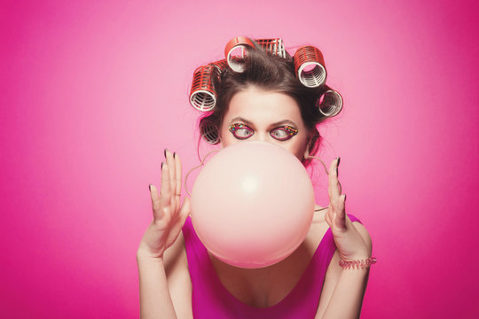 Cheeky girl with bubble gum posing on pink background in body, with curlers on head. Pretty woman with sweet makeup making balloons with bubblegum in studio