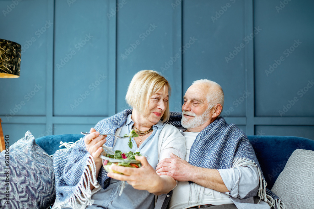 Wall mural Lovely senior couple eating healthy salad while sitting wrapped with plaid on the couch at home