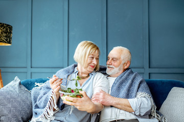 Lovely senior couple eating healthy salad while sitting wrapped with plaid on the couch at home