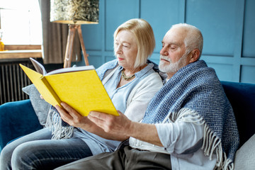 Lovely senior couple reading book while sitting wrapped with warm plaid on the couch at home