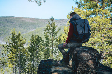 Man hiker with a large backpack sitting on a rock cliff and looking at the sprawling green valley. Freedom in travel