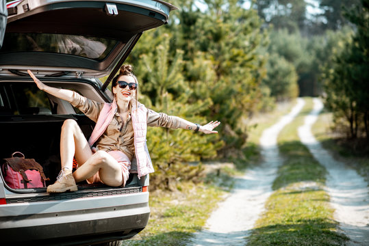 Young woman enjoying nature while sitting in the car trunk on a picturesque road in the woods