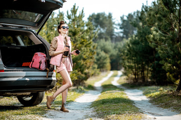 Young woman traveler enjoying the trip, standing with hot drink near the car trunk on a picturesque...
