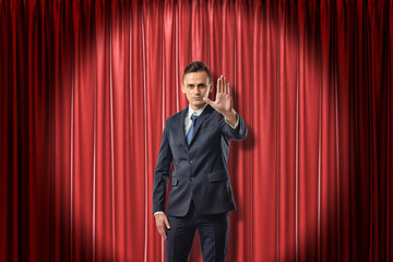 Front view of businessman standing in spotlight against red stage curtain looking at camera and holding out hand with open palm.