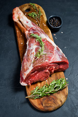 Raw leg of lamb on a wooden chopping Board. Rosemary, thyme, black pepper. Black background, top...