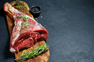 A raw leg of lamb on a wooden chopping Board. Rosemary, thyme, black pepper. Black background, side...