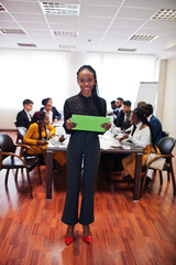 Face of handsome african business woman, holding clipboard on the background of business peoples multiracial team meeting, sitting in office table.
