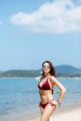 Beautiful brunette girl in a red bikini swimsuit and sunglasses posing on the beach on a Sunny day. Portrait of a woman on vacation against the sea.