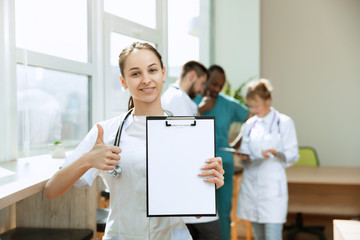 Beautiful smiling female professional doctor standing over hospital background. Caucasian woman showing empty banner with copyspace. Advertising image about healthcare, health, clinic, medicine.