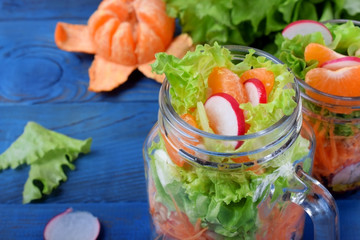 Layered salad in a jar with mandarins, radish, lettuce leaves, carrot and dressing against the blue background