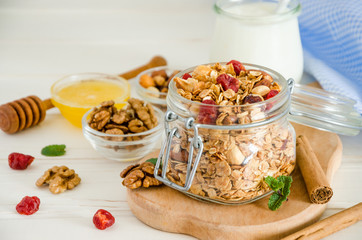 Multicereal homemade organic granola with a mixture of nuts, dried cherries, honey, cinnamon  and yogurt in a glass jar on a white wooden background.