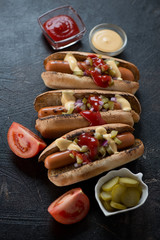 Hot-dogs with grilled sausages, pickles, red onion and dipping sauces, studio shot over dark brown stone background