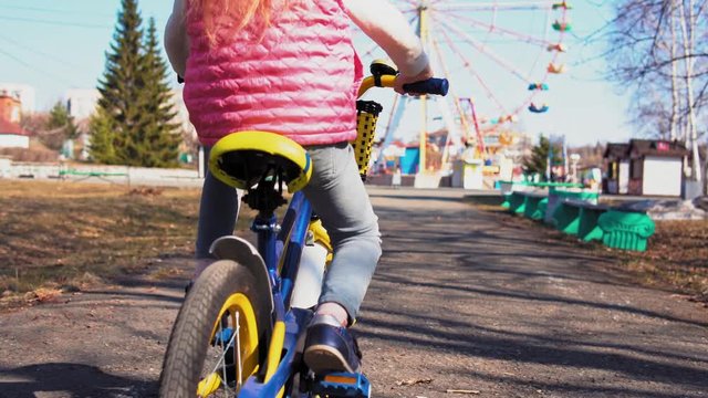 Handheld back view shot of five-year-old little blonde girl in red protective helmet ride bike with training wheels on asphalt track with gaps and holes sunny day in old park.