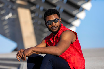 Front view of a young black man wearing sunglasses sitting on staircase in a sunny day while looking away