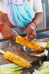 Close-up hands of female street vendor is rubbing a roasted sweet corn cob with lemon and spices. Indian street food concept, closeup
