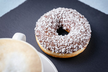 Chocolate-covered doughnut and coconut shavings on a slate next to a Cup of cappuccino.