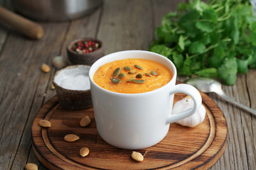 Pupmkin cream soup puree in cup on brown wooden table, side view. Dietary vegetarian lunch on cutting board with parsley, garlic.