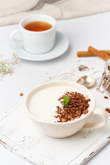 Yogurt with chocolate granola in cup, breakfast with tea on white wooden background, vertical.