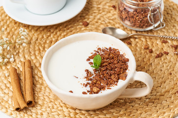 Yogurt with chocolate granola in cup, breakfast with tea on beige background, side view, close up.