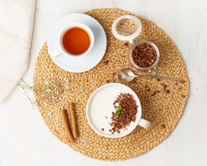 Obraz na płótnie Canvas Yogurt with chocolate granola in cup, breakfast with tea on beige background, top view.