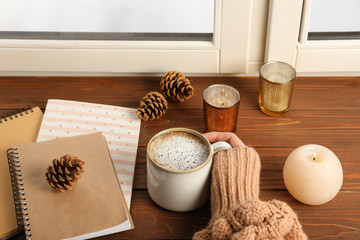 Woman with cup of coffee and notebooks at window indoors, closeup. Winter drink