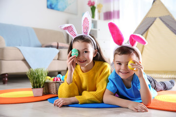 Cute children with bunny headbands and painted Easter eggs lying on floor at home