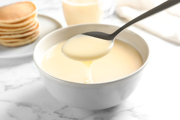 Spoon of pouring condensed milk over bowl on marble table, closeup. Dairy products