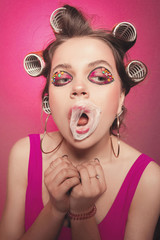 Cheeky girl with bubble gum posing on pink background in body, with curlers on head. Pretty woman with sweet makeup making balloons with bubblegum in studio. Balloon burst on lips