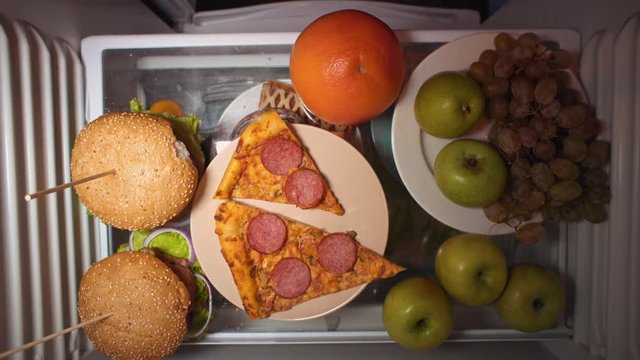 Top view on the shelf of the refrigerator. A man's hand is trying to take a hamburger, but women's fingers are directed to the grayfruit.