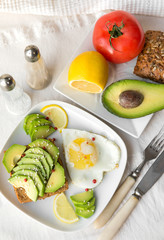 sandwich with avocado, sesame and eggs, half avocado, tomato, lemon , bread, fork, knife, salt ,pepper on a white tablecloth