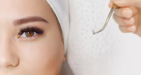 Eyelash Extension Procedure. Close up view of female brown eyes with long eyelashes. Stylist holding tweezers, tongs and making lengthening lashes. Macro, selective focus.