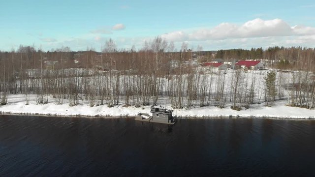 Aerial, drone shot, around a foating sauna, behind leafless trees, on a river, on a sunny, winter day, in Marjala, near Joensuu, North Karelia, Finland