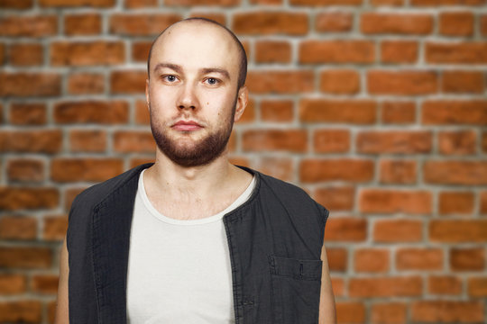 Portrait Young bald guy with beard with pocker face dressed in sleeveless shirt. Man on brick. wall background.