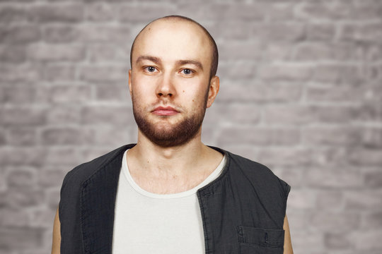 Portrait Young bald guy with beard with pocker face dressed in sleeveless shirt. Man on brick wall background.