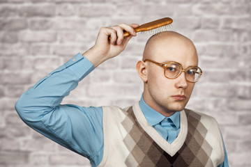 Portrait of a bald guy in a blue shirt holding a comb in his hands on brick wall white background....
