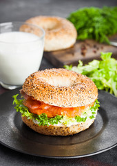 Fresh healthy bagel sandwich with salmon, ricotta and lettuce on black plate on black kitchen table background. Healthy diet food. Glass of milk and fresh vegetables