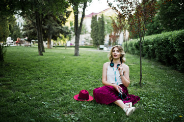 Fashionable and beautiful blonde model girl in stylish red velvet velour skirt, white blouse and hat, sitting on green grass at park with phone and earphones.
