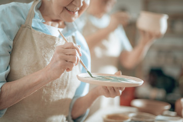 Cropped photo of elderly craftswoman painting decorative plate in workshop