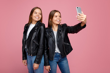 Beautiful two young twin sisters in leather jackets with bright smile use smartphone over pink background. Twin sisters take selfie with mobile phone.